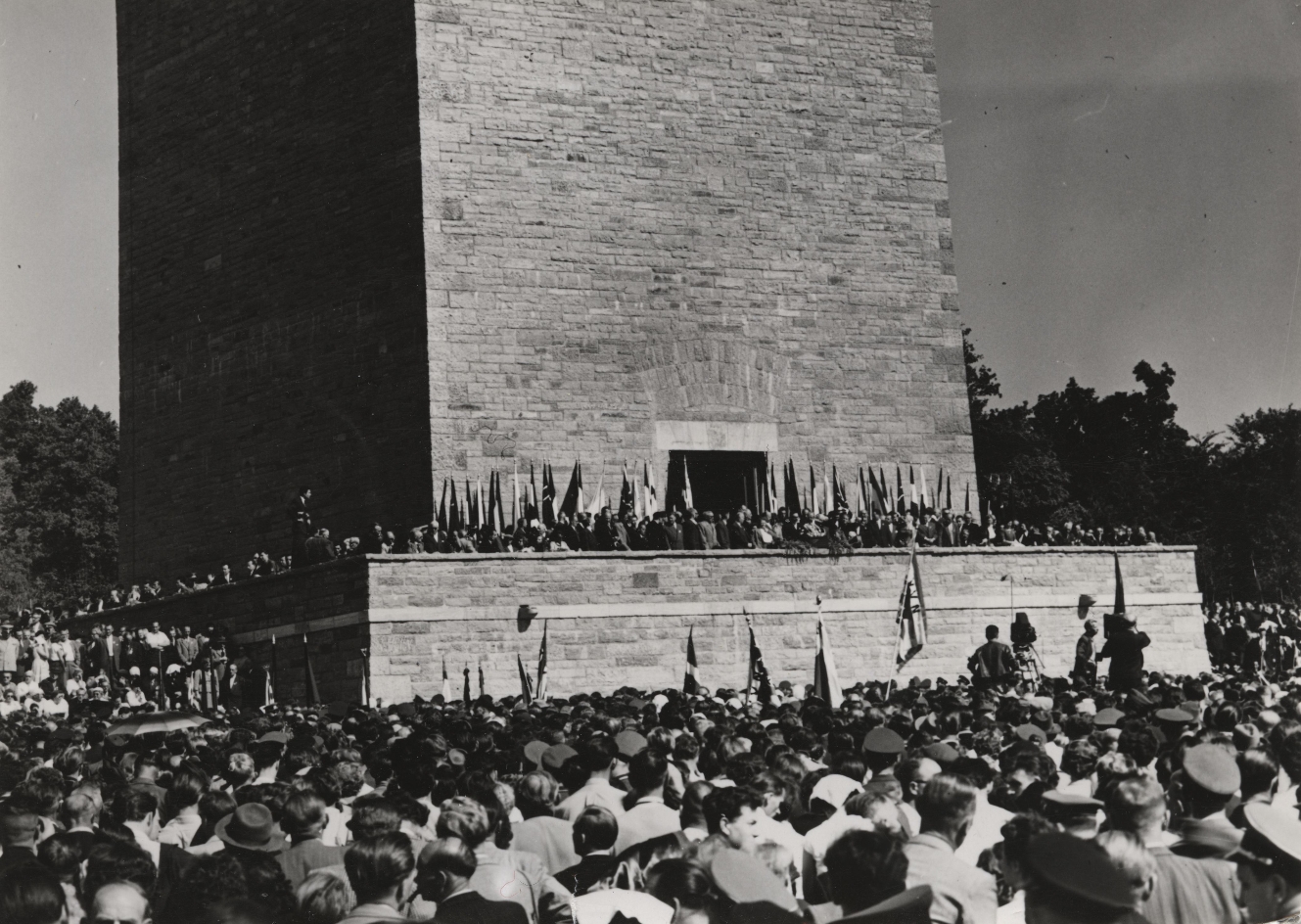 A large crowd of people with isolated flags standing in front of the Tower of Liberty and to another crowd with higher density of flags at the entrance of the Bell Tower.