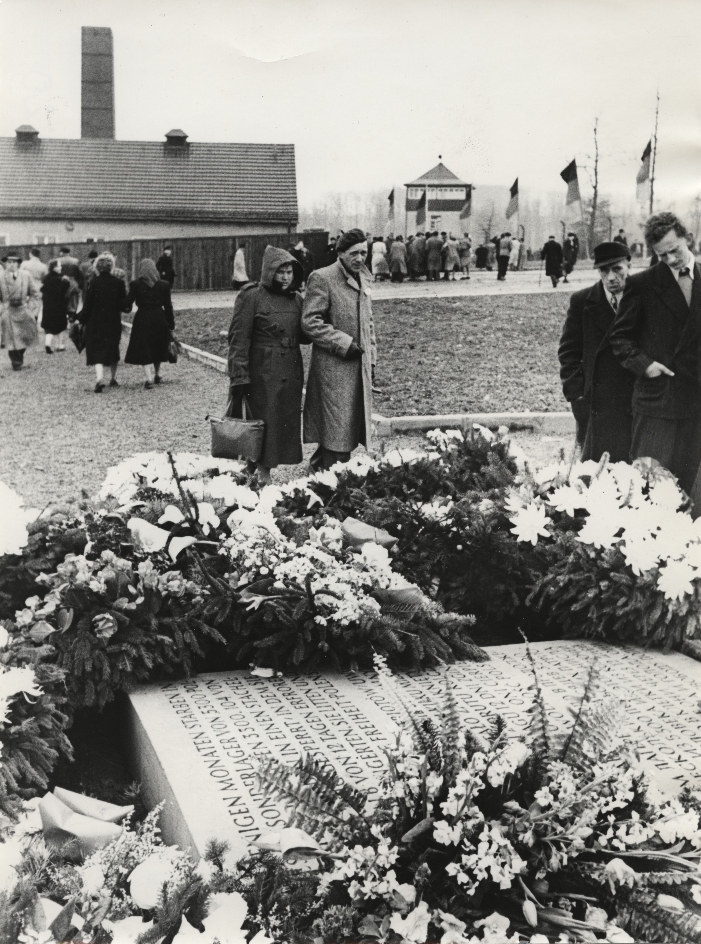 A memorial stone on the grounds of the former prisoners' camp, around which wreaths and flower arrangements have been laid. Passing visitors to the memorial site look concerned.