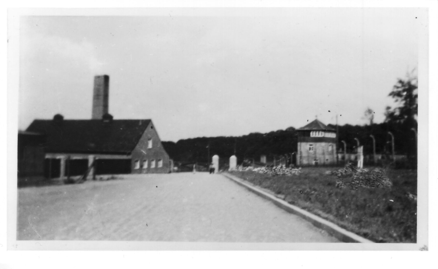 Unscharfes Bild einer Lagerstraße, die zum Krematorium führt. Rechts im Hintergrund ist ein Wachturm, links das Krematorium. Rechts auf dem Grasstreifen vor dem Lagerzaun erkennbare Spuren von Retuschen. 