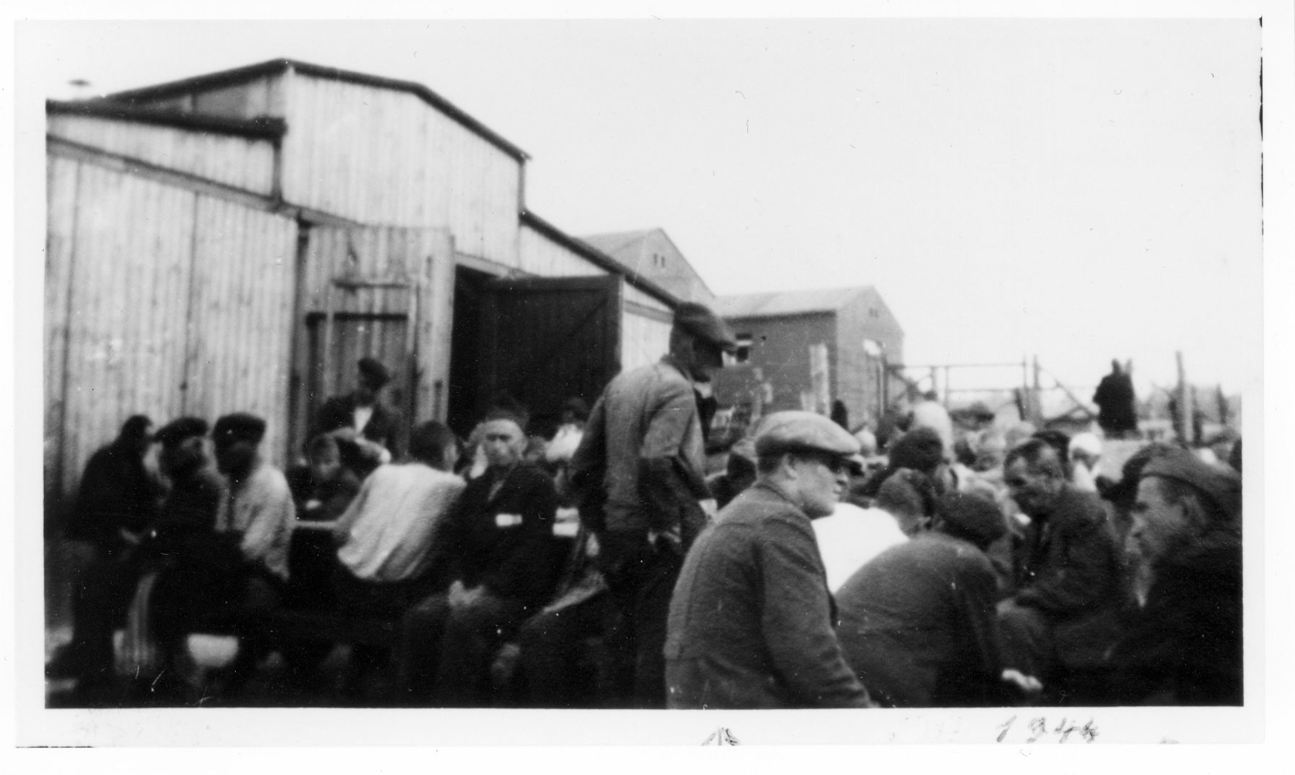 Häftlinge des Kleinen Lagers sitzen und stehen vor einer Pferdestallbaracke, dahinter zwei größere Steingebäude. Auf dem Bild ist ein geschäftiges Treiben zu erkennen. 