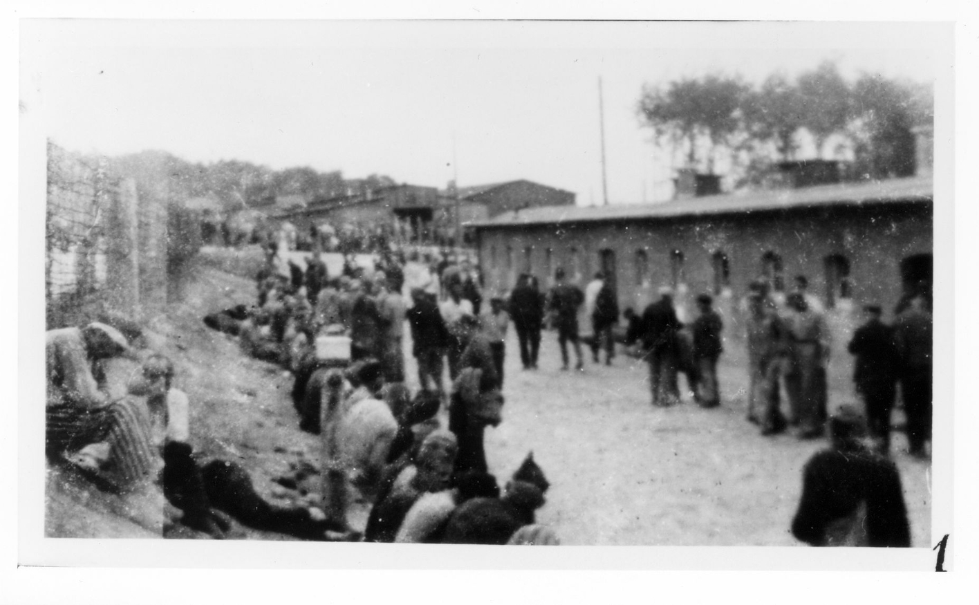 Prisoners on the camp road in front of the latrine and washroom building in the Small Camp. Barracks 59 and 62 in the background. Many prisoners are sitting by the roadside.