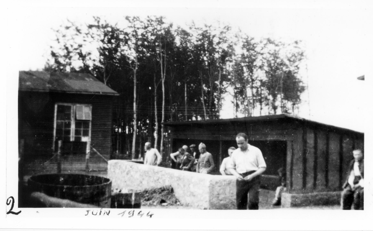 Häftlinge stehen an einer der kleinen Latrinen im Kleinen Lager. Links im Bild die Kinobaracke. Handschriftlich "2" auf linkem unteren Bildrand und "JUIN 1944" auf unterem Bildrand
