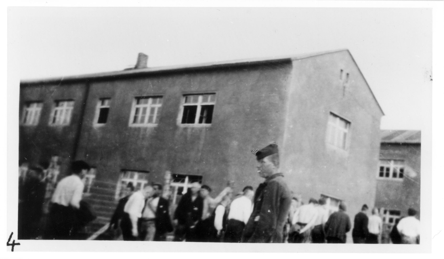 Häftlinge laufen vor einem der Steinblocks von links nach rechts. Die Mütze des im Vordergrund stehenden Häftlings ist retuschiert.