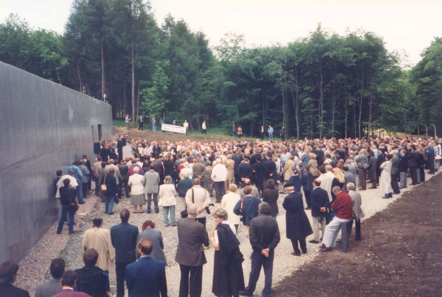 Vor einem grauen Gebäude am linken Rand befindet sich eine Ansammlung vieler Menschen auf einer Wiese und einem Weg. Im Hintergrund ist ein Waldstück zu sehen.