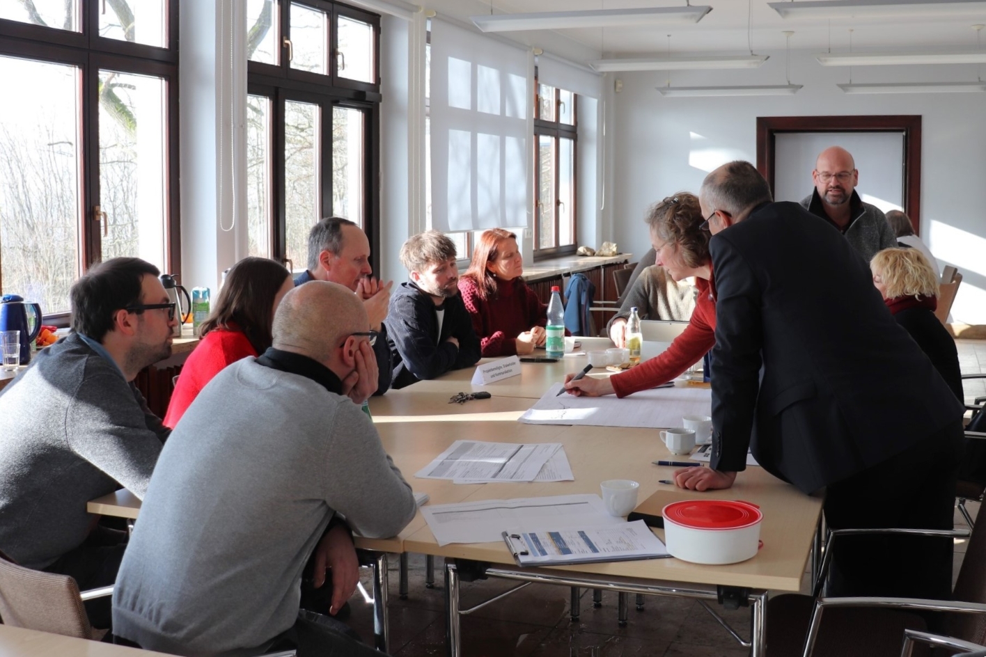 In einem langgezogenen Raum steht ein großer Konferenztisch. Menschen sitzen darum und hören zu. Rechts steht ein Mann im Vordegrund. Eine Frau hinter ihm erklärt etwas. Dahinter steht ein Mann.
