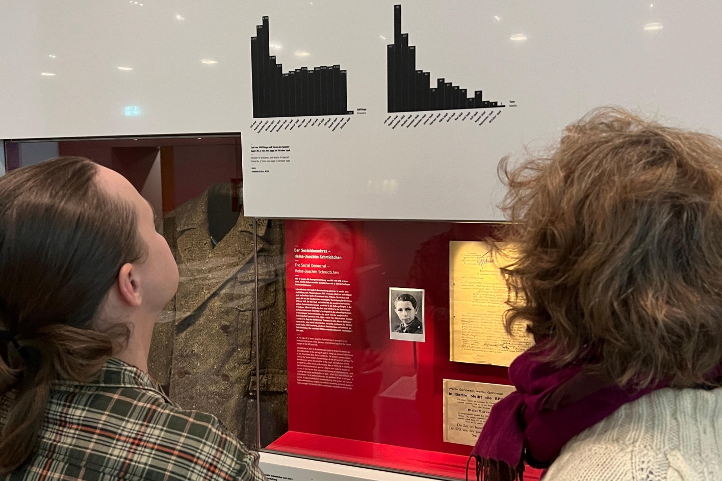 Kustodin Frau Dr. Julia Landau und Herr Vladislav Drilenko stehen sich in der Ausstellung der Gedenkstätte Hohenschönhausen vor einer Vitrine. Sie schauen sich eine Statistik über der Vitrine an. In der Vitrine hängt ein alter Mantel und ein Foto eines jungen Mannes. Es zeigt des Sozialdemokrat Hein-Joachim Schmidtchen.