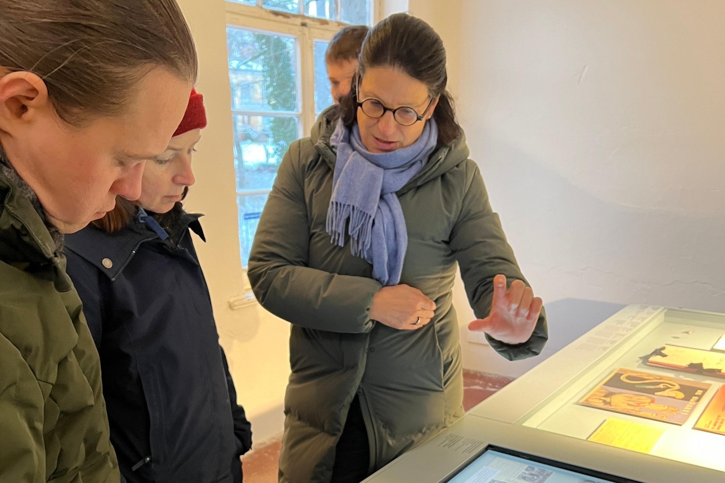 Rechts im Vordergrund ist ein Vitrinentisch zu sehen. Eine Frau, welche mittig im Bild zu sehen ist, erklärt etwas. Hinter ihr steht eine Person. Links neben ihr im Vordergrund steht eine Frau mit roter Mütze und ein junger Mann.
