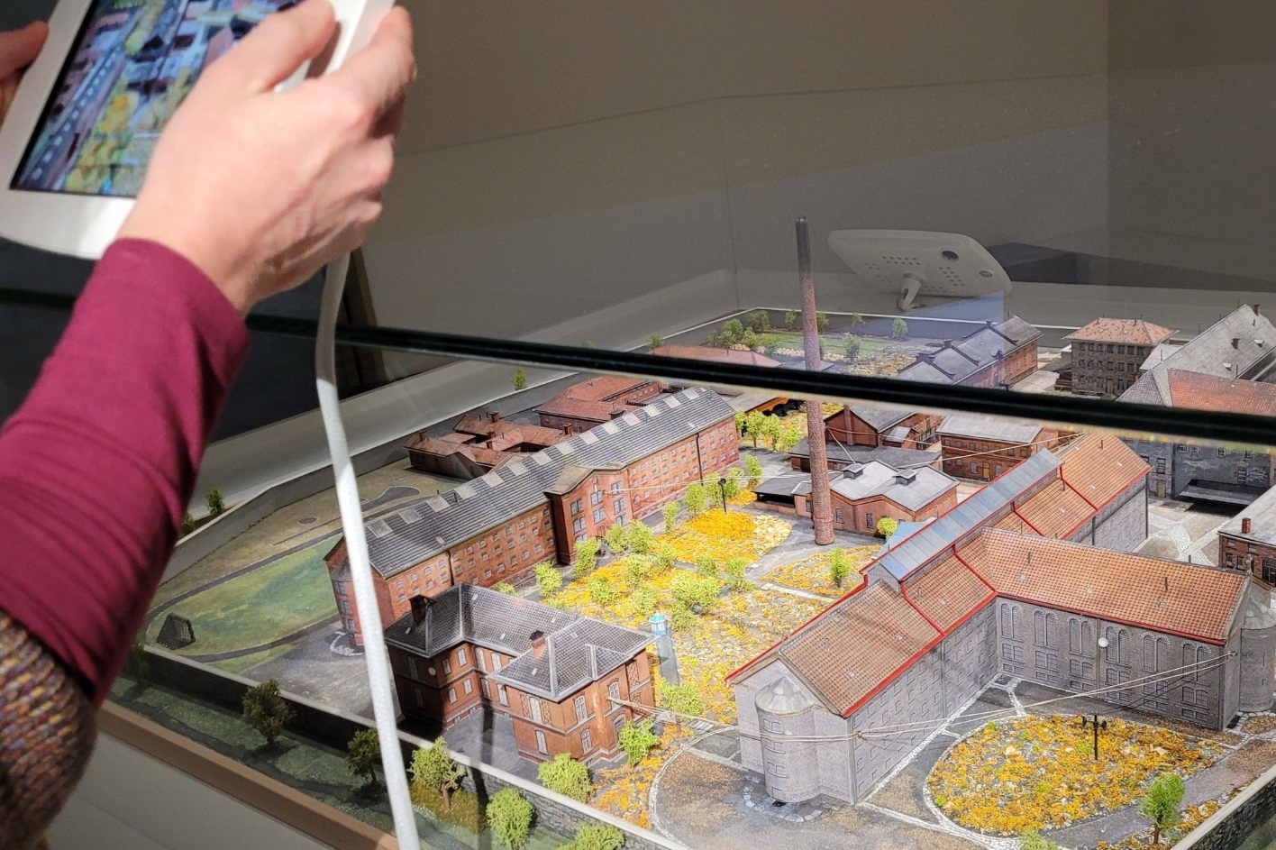 Eine Vitrine mit einem Model der Gedenkstätte. Eine Frau hält ein iPad in beiden Händen, welches auf das Model gerichtet ist.