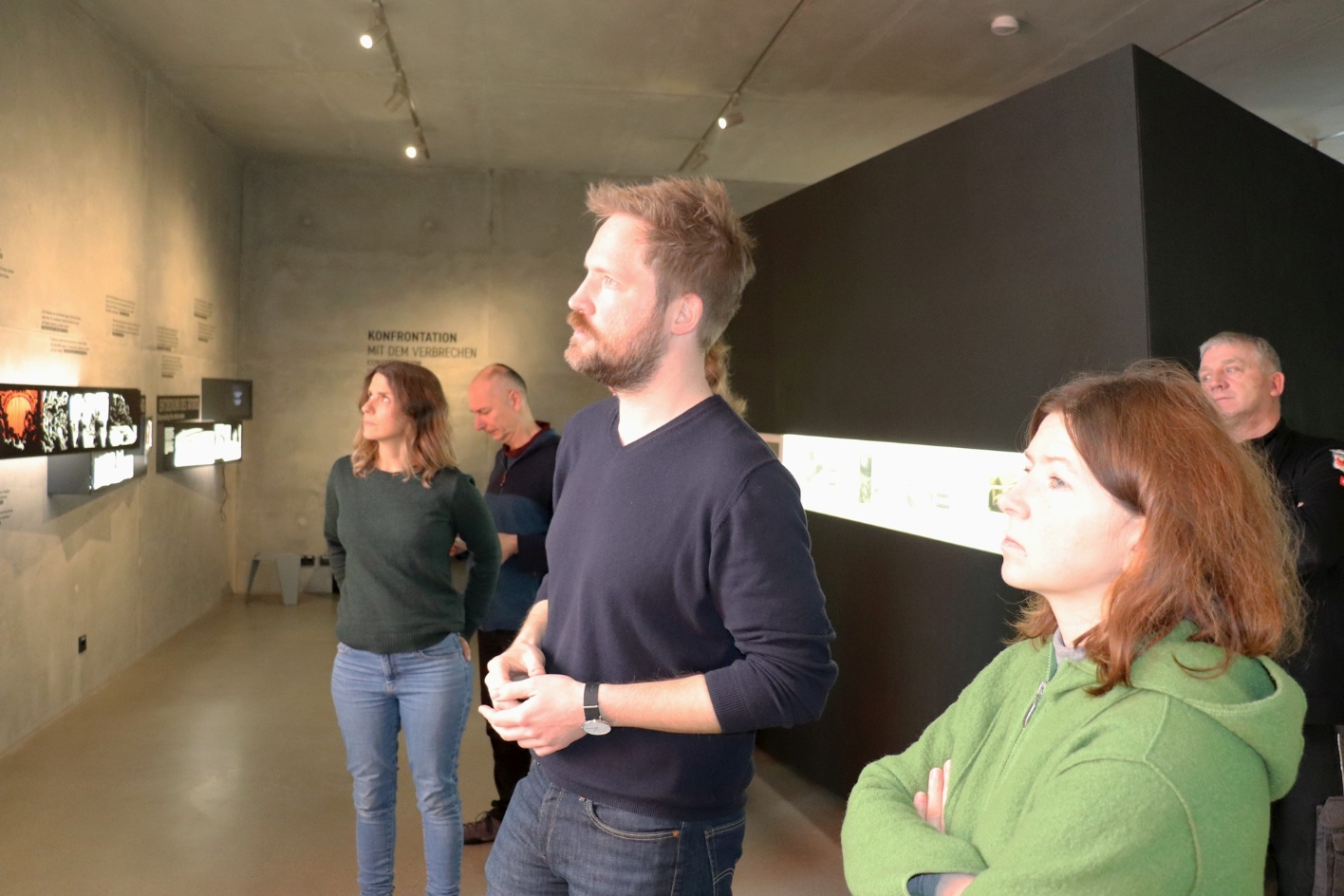Der kommissarische Leiter der Gedenkstätte, Lukkas Busche, erläutert dem Team die Ausstellung. Zu sehen sind einzelne Ausstellungselemente auf grauer Betonwand.