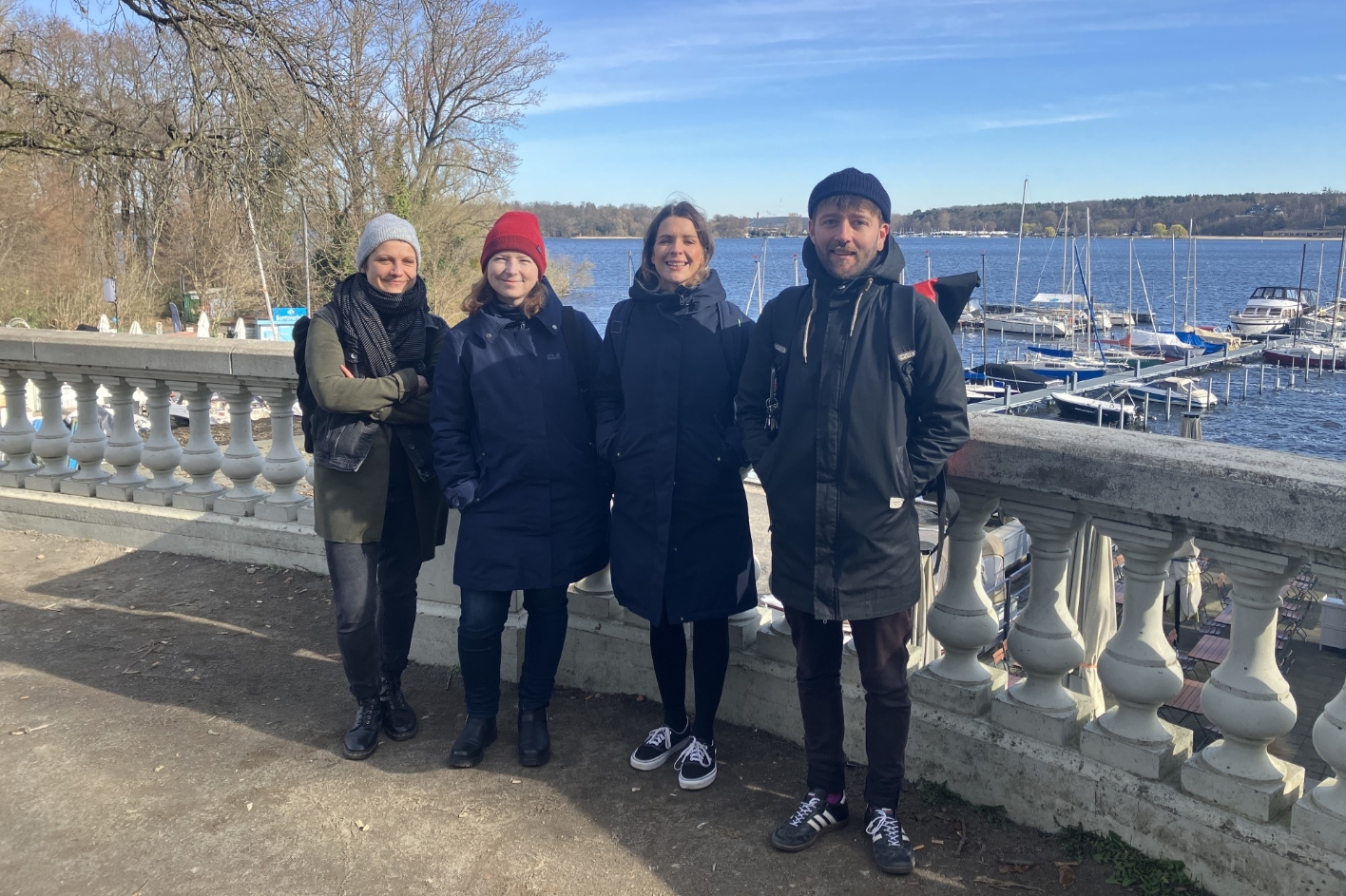 Im Vordergrund stehen drei weibliche Personen und eine männliche Person nebeneinander vor einem Geländer. Im Hintergrund sind Schiffe auf einem See zu sehen.