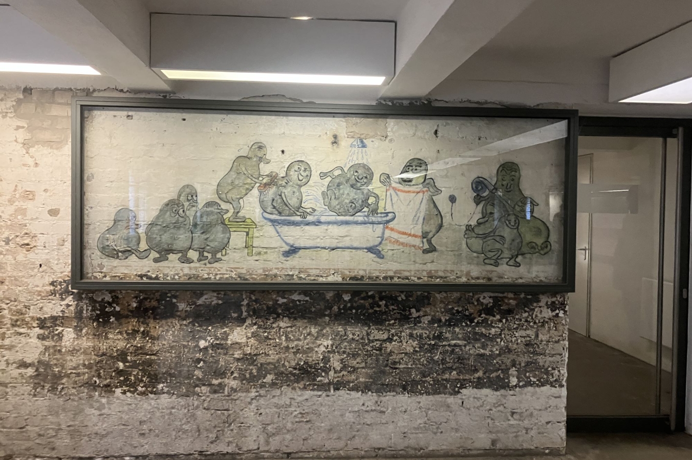 Zu sehen ist eine brüchige Steinwand, auf der sich eine Malerei befindet, die zum Schutz von einem Glaskasten eingerahmt ist. Die Malerei zeigt Kartoffeln mit menschlichen Gesichtern, die ein Bad nehmen. Andere Kartoffeln stehen um sie herum. Eine davon hält ein Handtuch bereit.