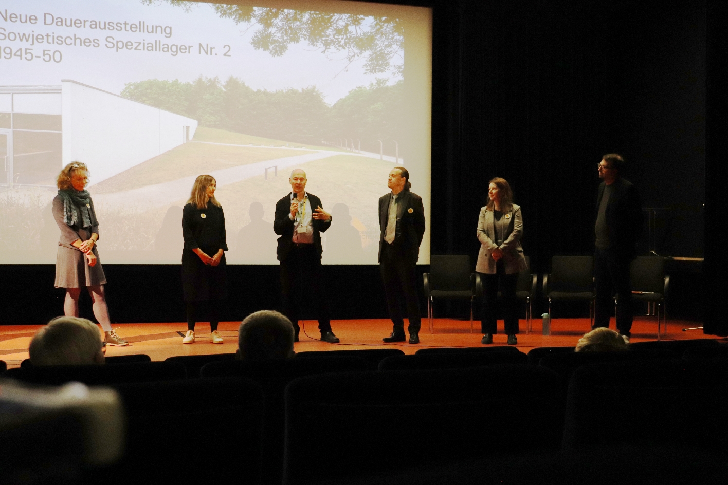 Auf der Bühne des abgedunkelten Kinosaals stehen fünf Personen. In ihrem Rücken ist eine Videoprojektion zu sehen. Auf ihr ist ein eingeschossiger Betonbau am Waldesrand zu sehen; als Titel steht auf der Präsentation: „Neue Dauerausstellung Sowjetisches Speziallager Nr. 2 1945 – 50“