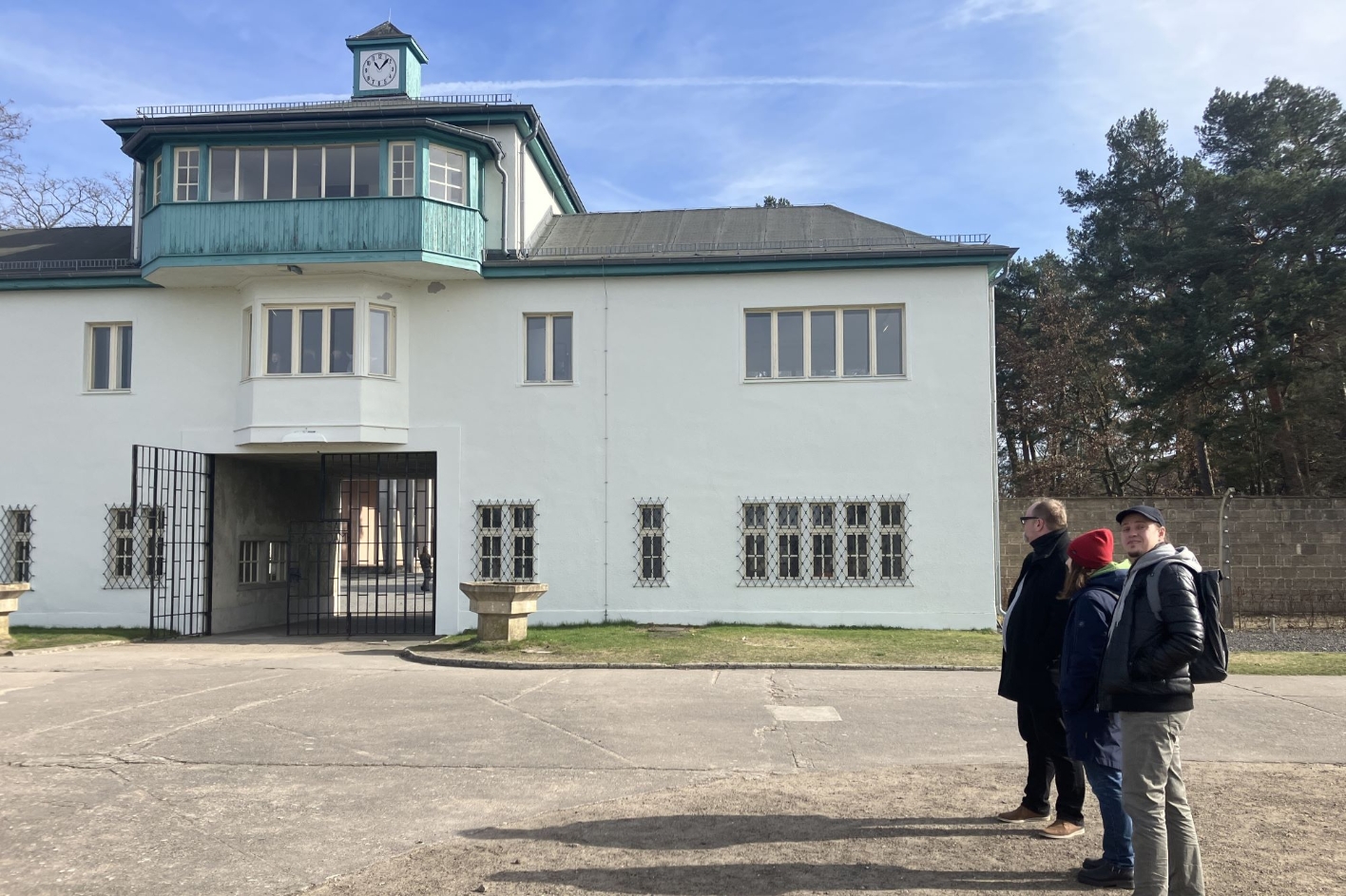 Zu sehen ist das große weiße Lagertor mit Aufbau der Gedenkstätte Sachsenhausen. Rechts davon stehen zwei Männer und eine Frau, die einen Blick auf das gebäude werfen.