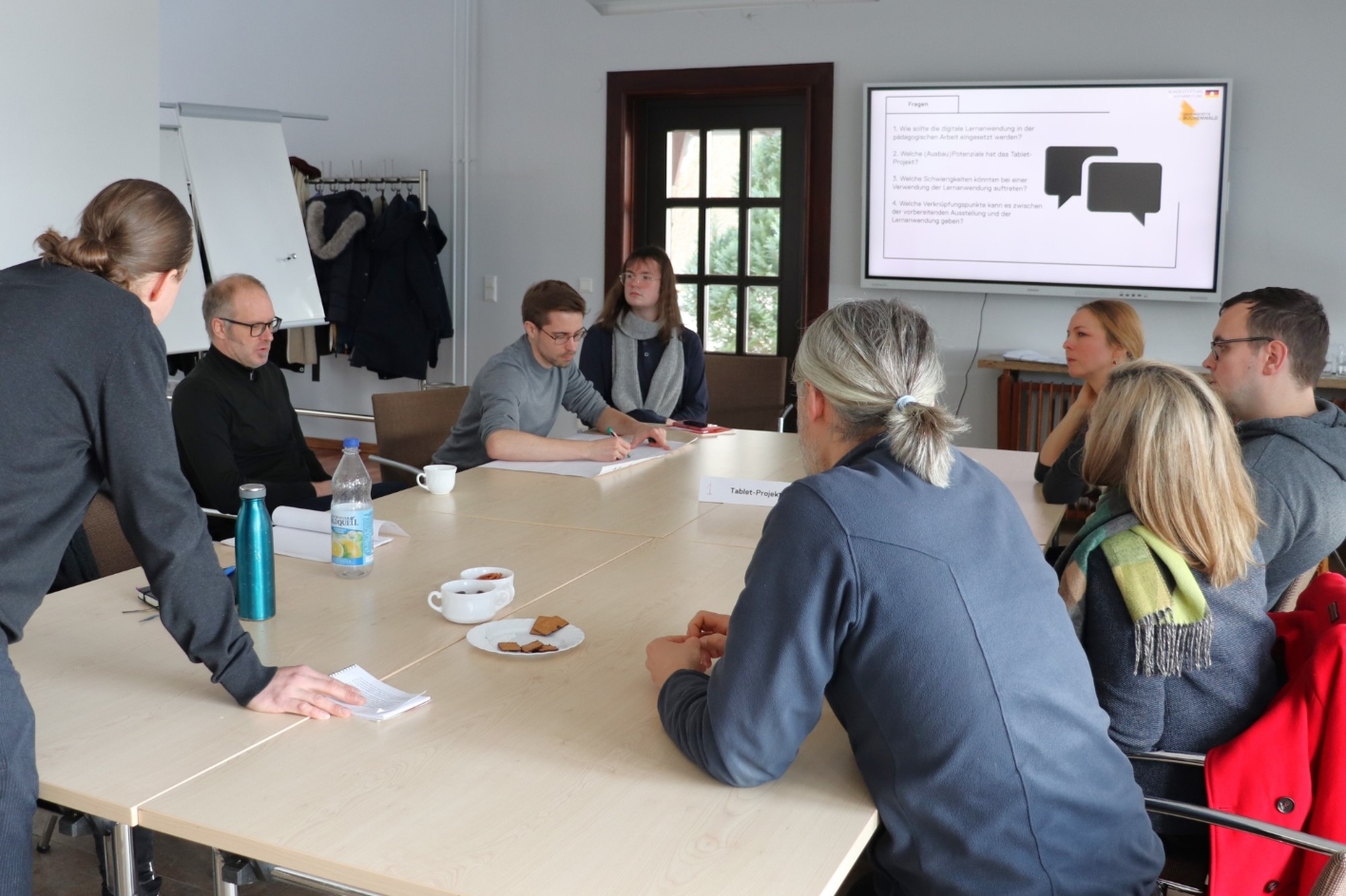 in der Mitte des Raumes steht ein Konferenztisch. Im Vordergrund steht an der Stirnseite ein Mann. Links und rechts sitzen Menschen. Im Hintergrund hängt eine elektronische Tafel.