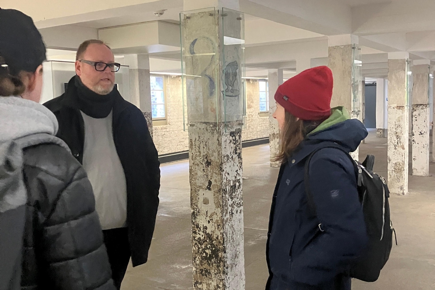 Auf dem Bild sind zwei Männer und eine Frau zu sehen, die gemeinsam in einem Raum mit vielen Säulen stehen. Ein Mann ist von vorne zu sehen und erläutert der Frau und dem anderen Mann die Räumlichkeiten.