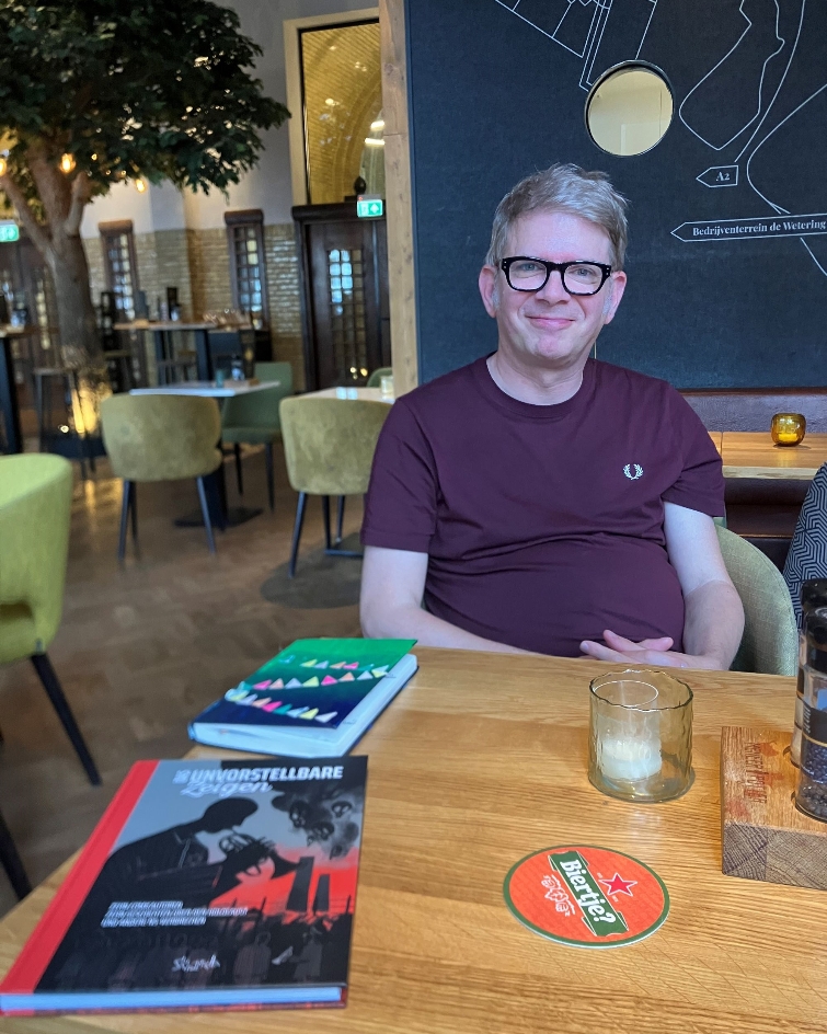 Ein Mann sitzt in einem Café am Tisch und lächelt in die Kamera. Auf dem Tisch liegen verschiedene Bücher, über die er spricht.