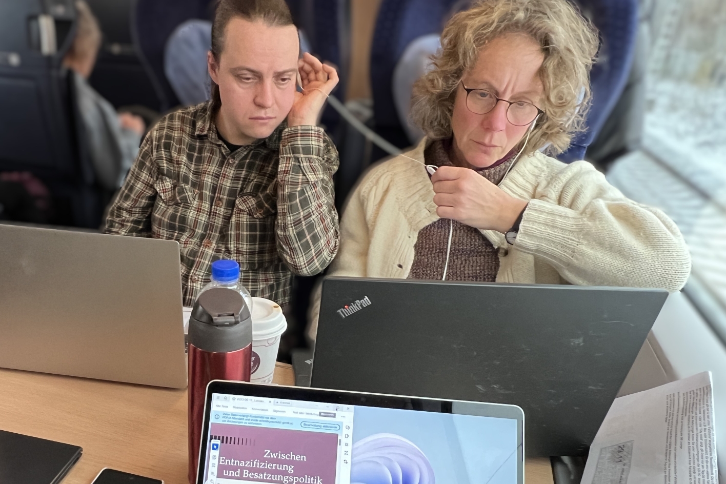 Kustodin Frau Dr. Julia Landau und Herr Vladislav Drilenko sitzen im Zug an einem Tisch. Vor ihnen stehen drei Laptops. Sie schauen auf den Bildschrim und haben Kopfhörer im Ohr.