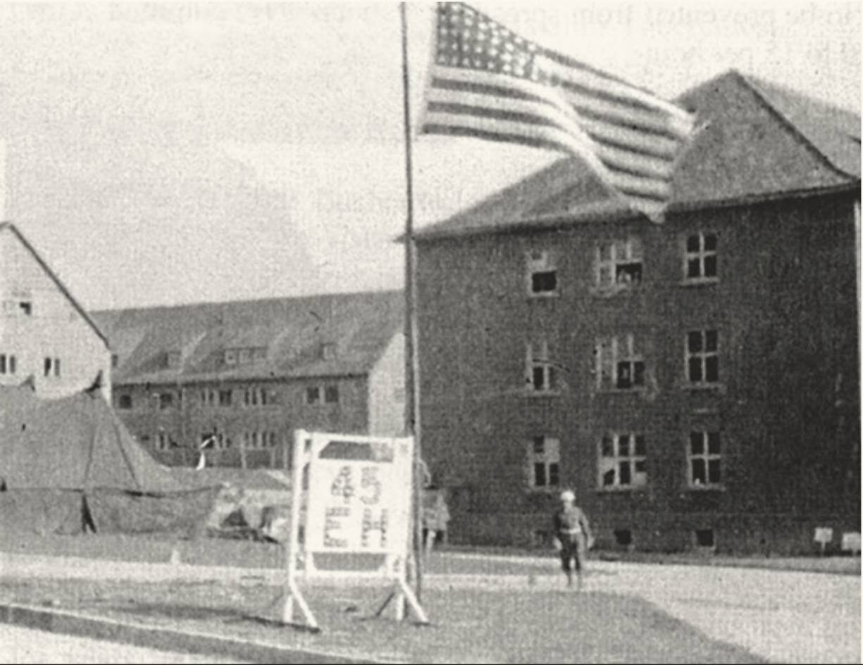 The image depicts a historical scene featuring an American flag waving on a flagpole in the foreground. In front of the flag, there is a sign with writing on it, though the text is only partially legible. In the background, several large, multi-story brick buildings with broken or open windows can be seen, suggesting a military or war-related setting. To the left of the image, there are tents, indicating a temporary camp or provisional facility. A soldier or a person in uniform is walking toward the camera.