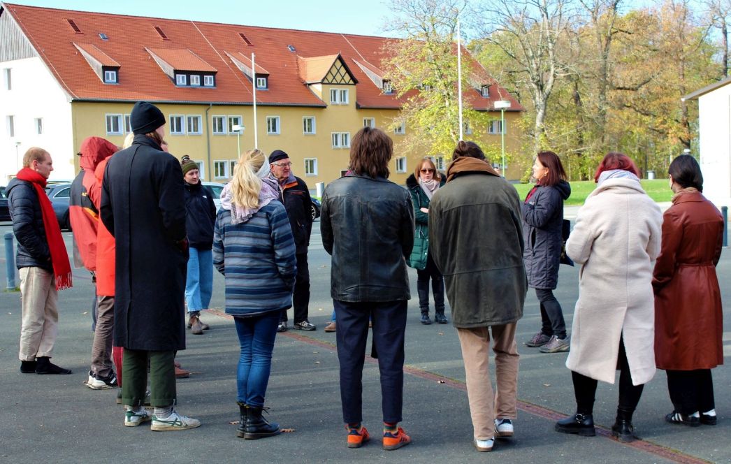 A group stands on the parking space. They are listening to a guide. In the background is a building of the International Youth Meeting Center.