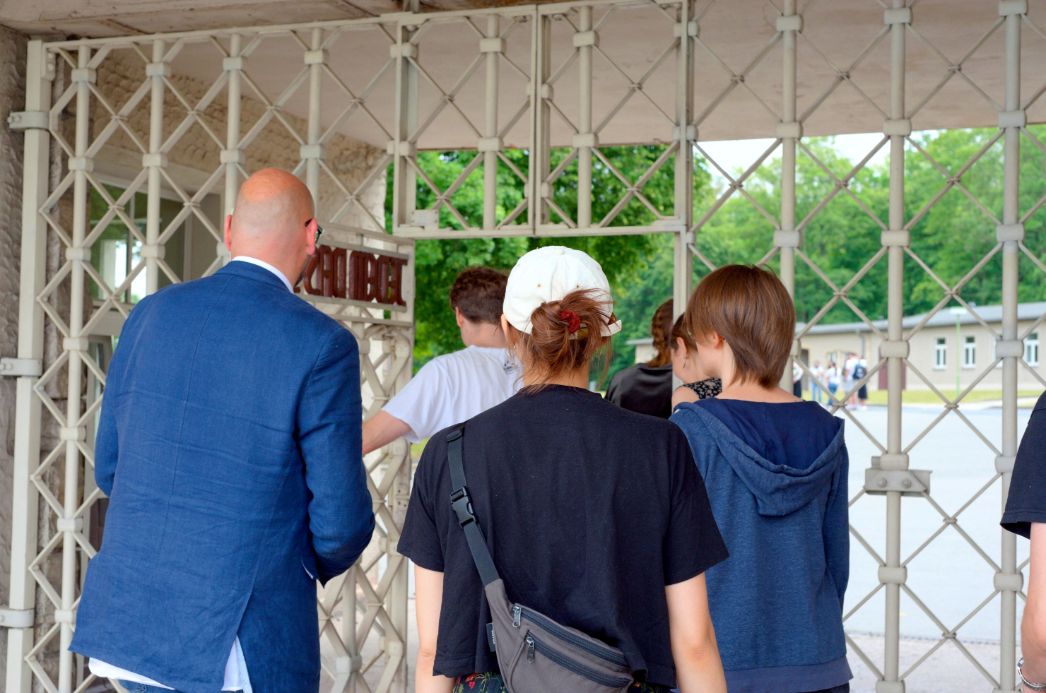 Einige Besucher mit ihrem Rundgangsleiter durchqueren das Lagertor. Links ist der Schriftzug "Jedem das seine" auf dem offenen Tor zu erkennen.