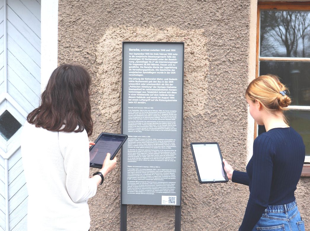 Zwei Teilnehmerinnen des Workshops stehen im Außenbereich der sowjetischen Verwaltungsbaracke vor einem Informationsschild. Sie beide haben Ipads in den Händen.