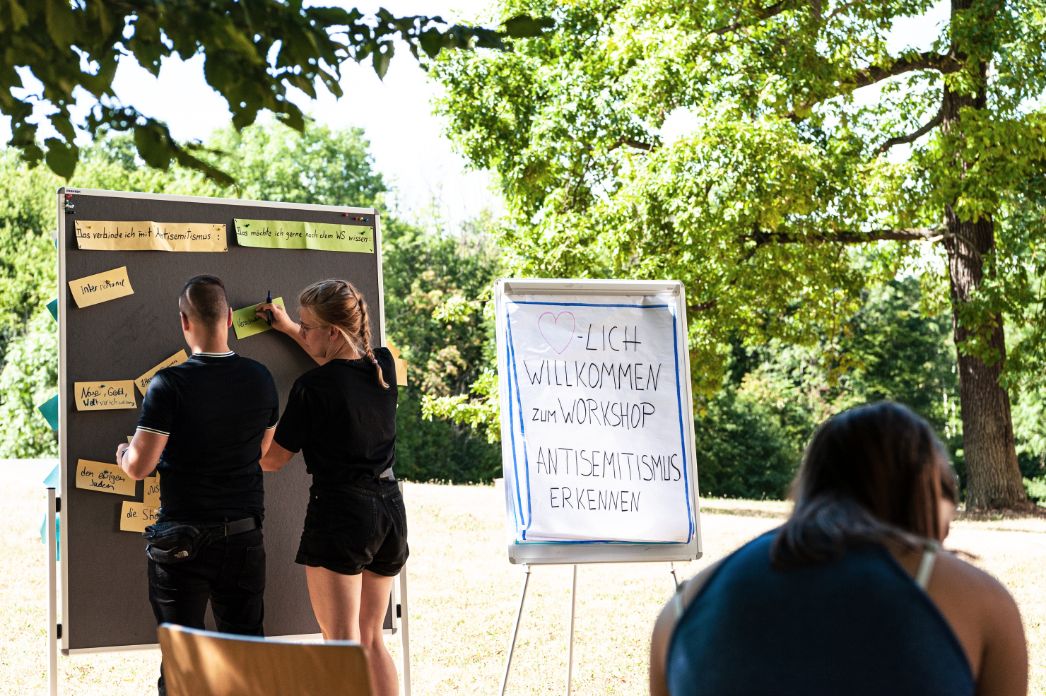 The picture shows 2 young people who are pinning something to a blackboard together. In the background on the right there is a poster with the inscription: "Welcome to the workshop "Recognizing anti-Semitism.