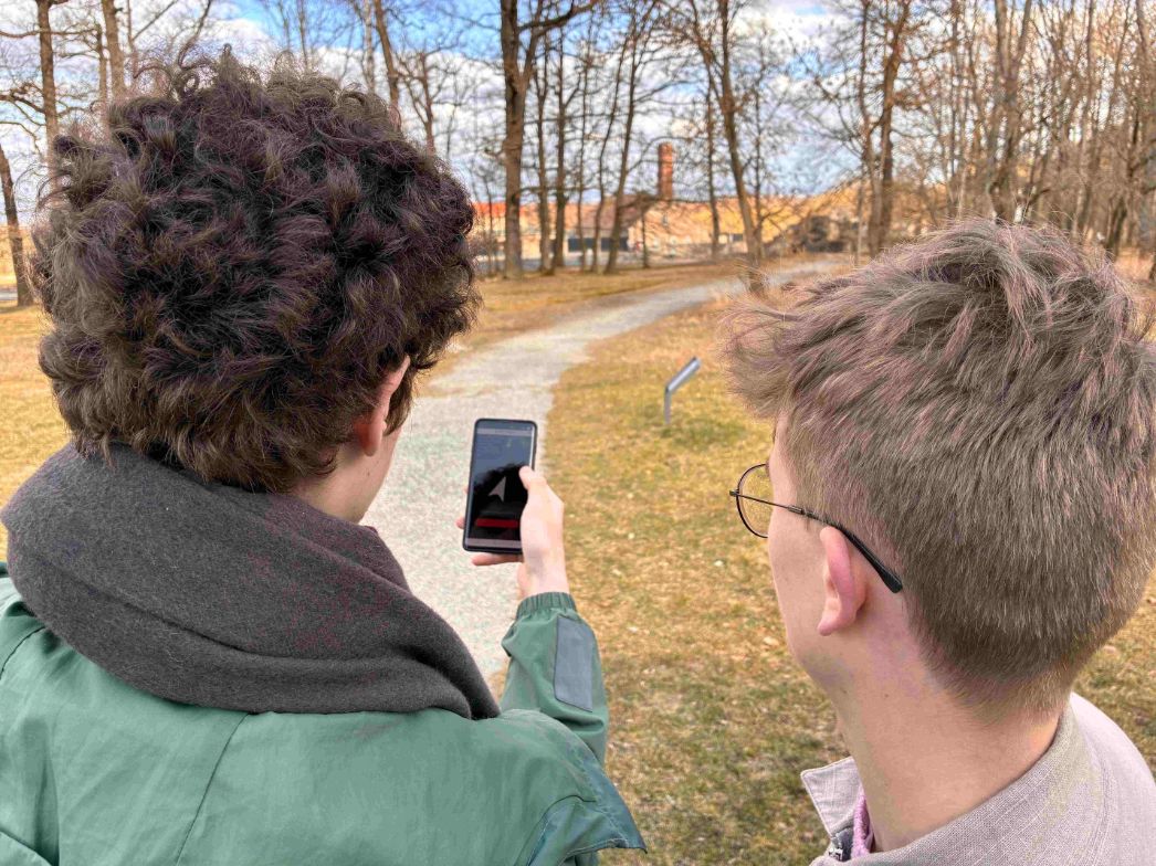 Zwei junge Schüler stehen in der Gedenkstätte am Rand des ehemaligen Zoos. Sie schauen auf ein Handy, das einer der beiden hochhält. Auf dem Handy ist ein Navigationspfeil zu erkennen, der ihnen die Richtung weist. Im Hintergrund sieht man den Lagerzaun und den Schornstein des Krematoriums.