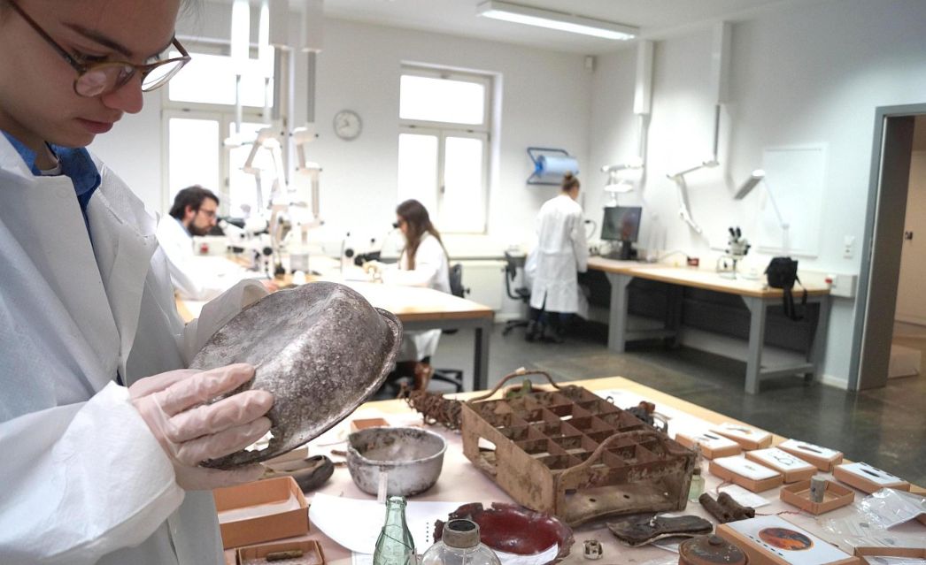 Eine Person reinigt eine alte Essschüssel. Auf dem Tisch vor der Person sind historische Objekte zu sehen. Menschen im Laborkittel im Hintergrund an Geräten.