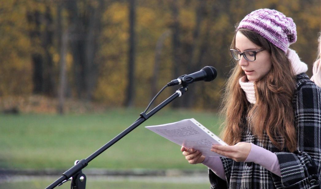 Eine junge Frau steht auf einer Wiese vor einem Mikrofon und ließt von einem Zettel ab.