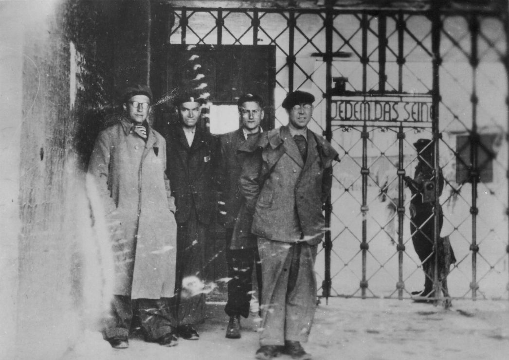Hermann Brill und drei weitere Männer stehen vor der nach innen angebrachten Lagertor-Inschrift „Jedem das Seine“.
