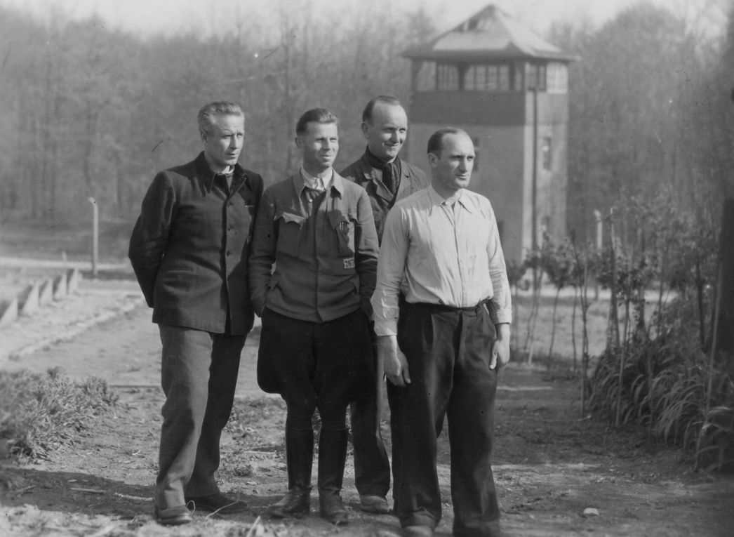 Group photo of four men including Otto Leischnig. In the background you can see a Buchenwald watchtower.