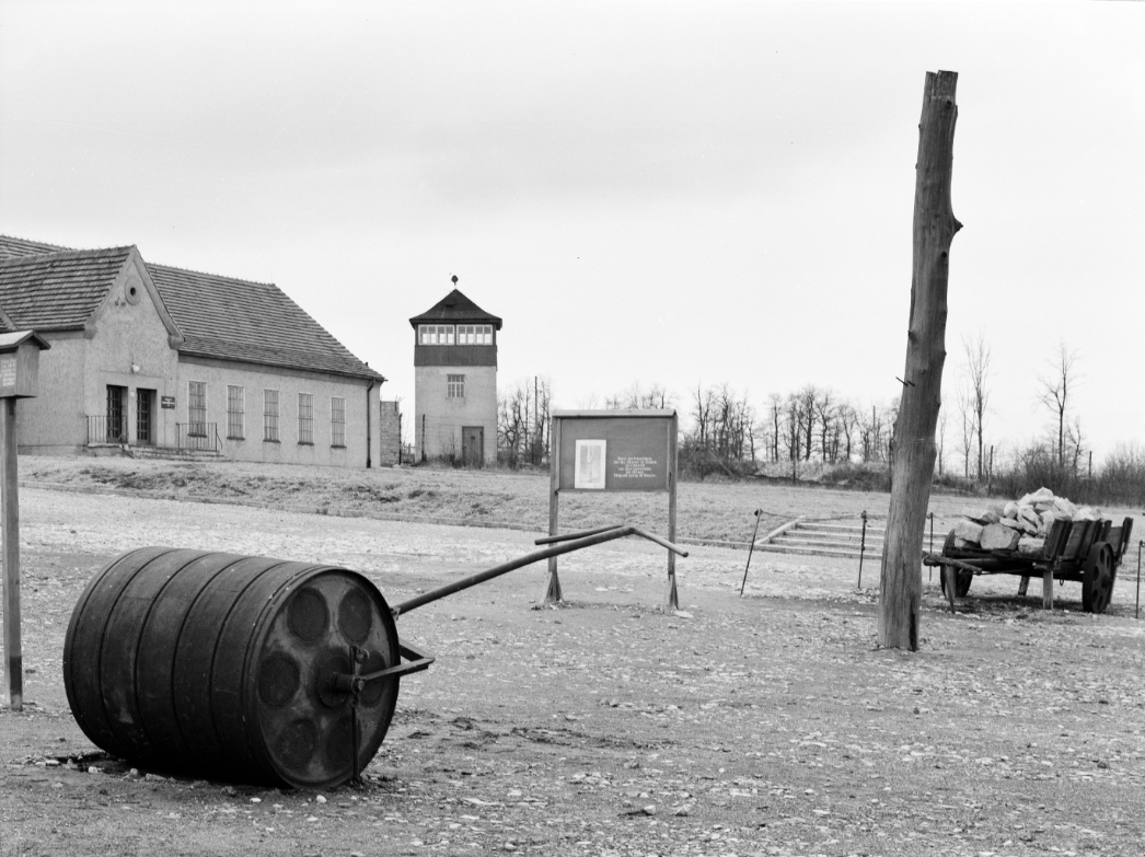 In the foreground, on the left side, is a hand leveler. A pile can be seen on the right. A little behind the pile is a cart loaded with quarry stones. In the center of the picture is a sign offering information to the visitor. In the left background, the entrance and right wing of the former prisoners' canteen can be seen. To the right is one of the watchtowers of the former camp.