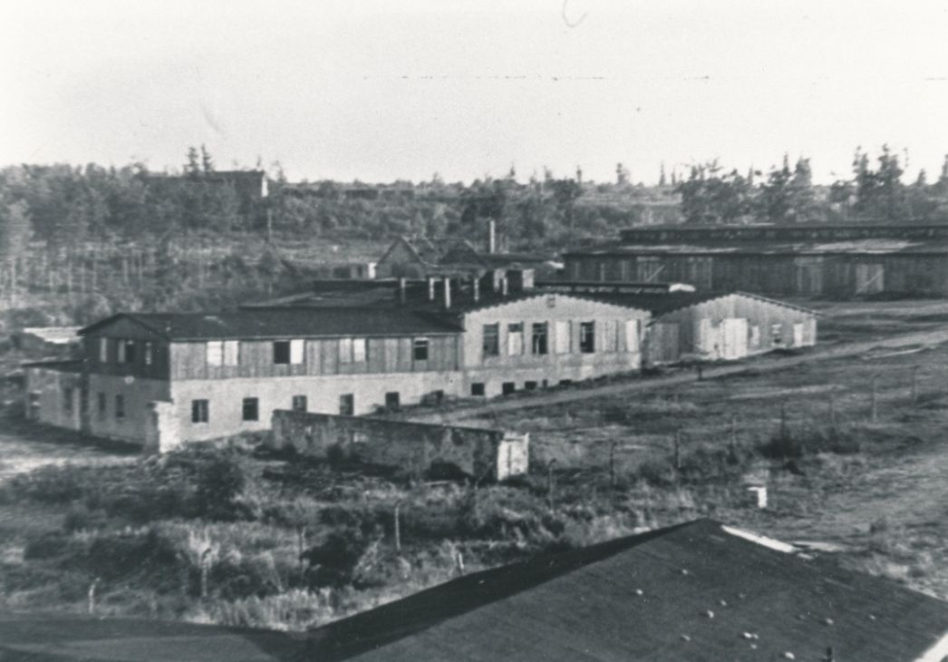 The picture shows the former workshop area after the dissolution of the special camp. In the center of the picture, a long, multi-story building can be seen. In the background a horse stable-like barrack. The complex is surrounded by thinned out rows of trees. 