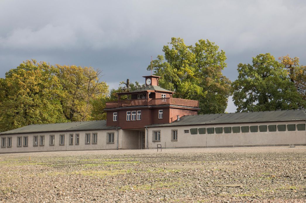Blick auf das Torgebäude vom ehemaligen Häftlingslagergelaende. Das Torgebäude hat zwei Flügel. In der Mitte des Gebauedes befindet sich der holzverschalte Hauptwachturm mit Lautsprecheranlage, Scheinwerfern und Uhr.