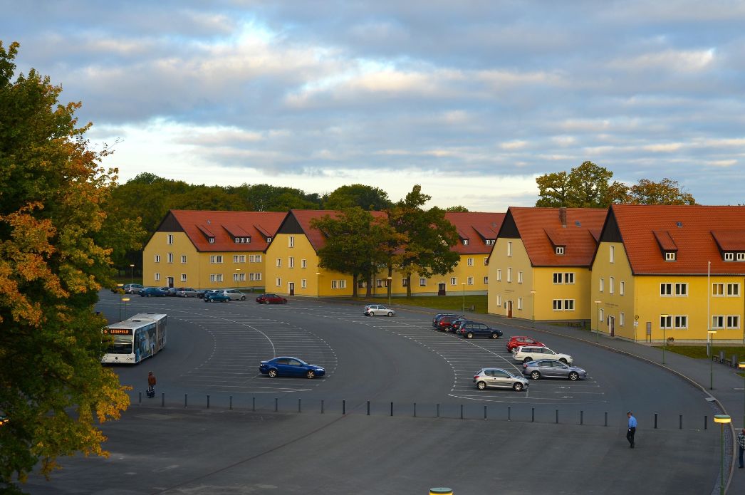 Blick auf den Parkplatz der Gedenkstaette. Links: Besucherinformation in einer der ehemaligen SS-Kasernen. Im-Hintergrund die ehemaligen SS-Kasernen, in denen heute die Jugendbegegnungsstätte angesiedelt ist.