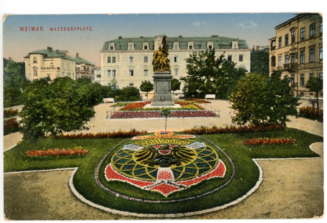 Es handelt sich um eine Postkartenmalerei. Zusehen ist ein Kriegsdenkmal auf der Mitte eines gestalteten Platz. Die gestelltung zeichnet sich durch aufwendig gepflegte Blumenbeete aus. Das Bild hebt die symetrische Gestaltung des Platzes nocheinmal hervor.