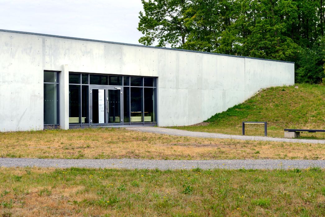 Das Foto zeigt den Eingang des Ausstellungsgebäudes von außen. DAs Gebäude hat ein flaches Dach. Daneben steht eine Bank.