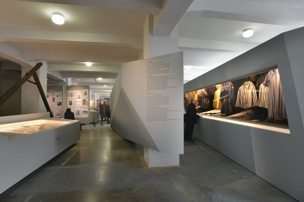 View into the exhibition. On the right, abstractly shaped showcases display items of clothing, and on the left, an aisle with more showcases displaying documents and photographs. 