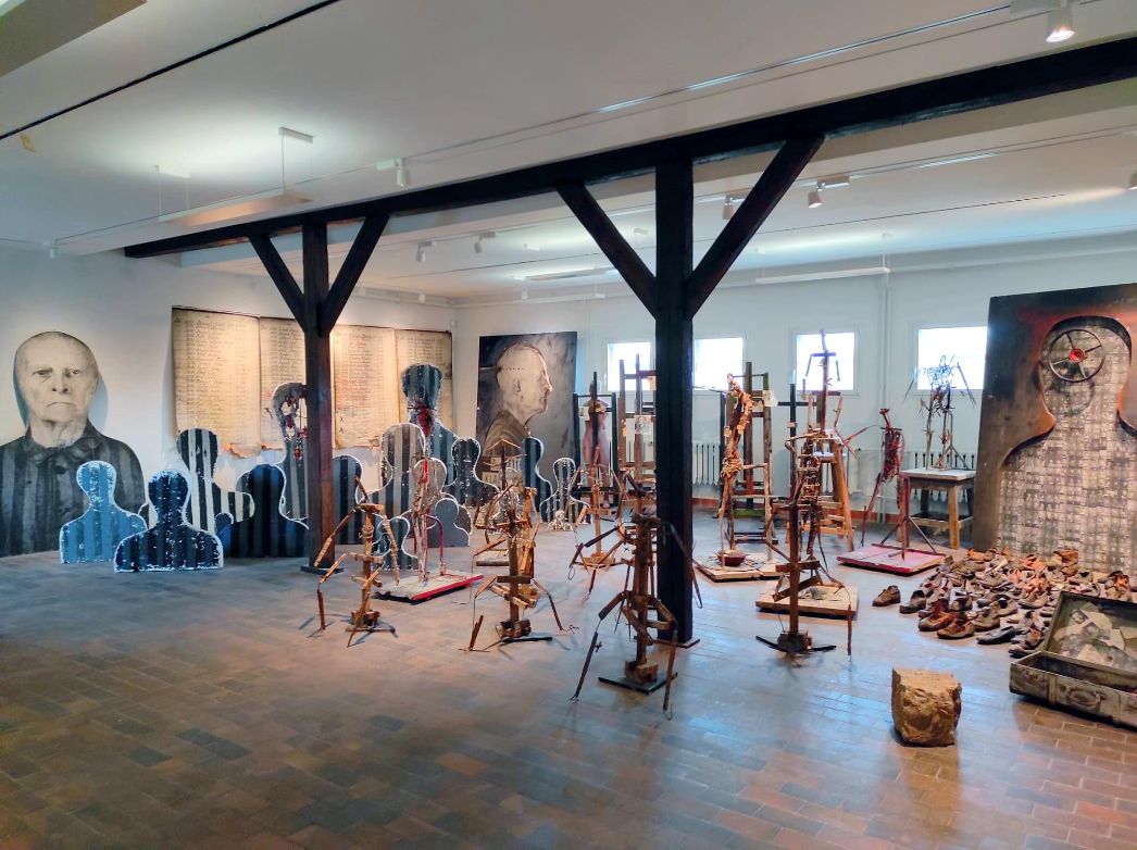 Blick auf die Installation Remineszenzen von Josef Szajna in der Kunstausstellung der Gedenkstätte Buchenwald. Dürre, abstrakte Holzgestelle füllen den Raum, sie erinnern vage an Menschen. Im Hintergrund überlebensgroße Porträts, eine große Liste, und Schuhe auf dem Boden.
