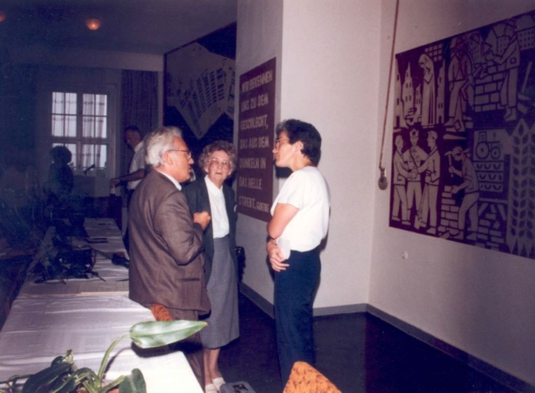 Drei Menschen stehen in einem Raum und diskutieren. Die beiden links und in der Mitte stehenden Personen sind älter. Im Raum befinden sich ein langer Tisch, ein Fenster und zwei Wandbilder.