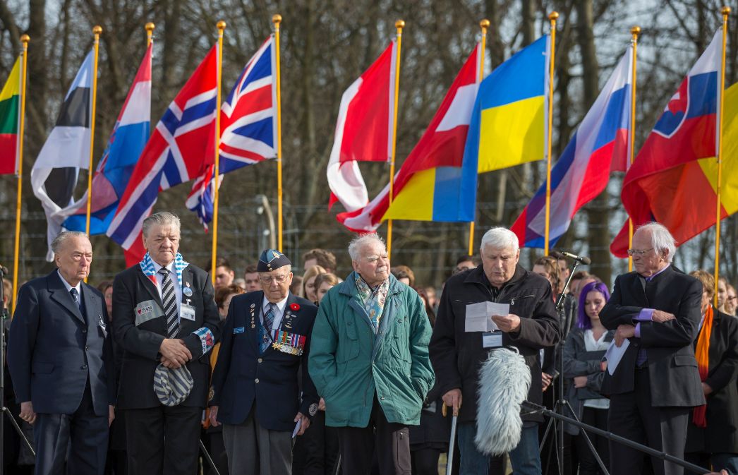 Mehrere ältere Männer stehen vor einer Menschenmenge, die Flaggen hochhält. Einer hat einen Zettel in der Hand und spricht. Einer trägt eine Uniform mit Abzeichen, einer hat einen roten Winkel und eine Nummer auf der Jacke. 