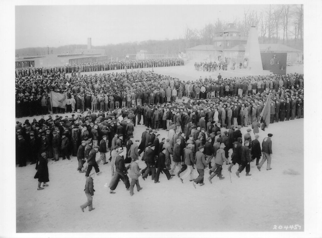 Befreite Häftlinge versammeln sich zum Gedenken an die Toten auf dem Appellplatz, 19. April 1945. Ein hölzerner Obelisk mit der Inschrift „K.L.B. 51.000“ (für die damals geschätzte Zahl der Opfer) diente als provisorisches Denkmal. Das Foto ist die einzige bekannte Aufnahme der Gedenkfeier. Foto: Donald R. Ornitz (U.S. Signal Corps)