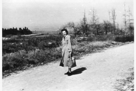A woman in civilian clothes stands on a path between single trees in barren landscape and smiles into the camera. 