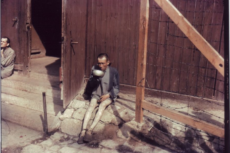 One color picture shows a liberated prisoner sitting alone in front of a barrack and looking at the photographer. He is extremely thin and holding a metal bowl.