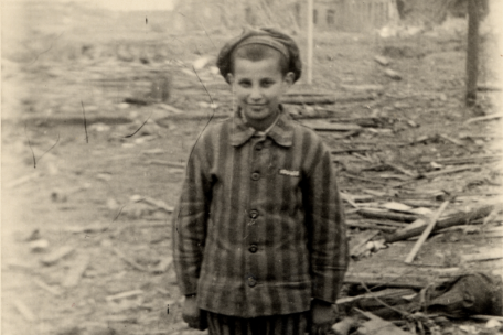 Ein Junge steht in Häftlingskleidung vor Trümmern und lächelt in die Kamera.