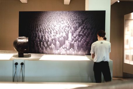 A young person at a listening station in the exhibition. In the center of the picture is a large print of a historical photograph depicting prisoners lined up for roll call.