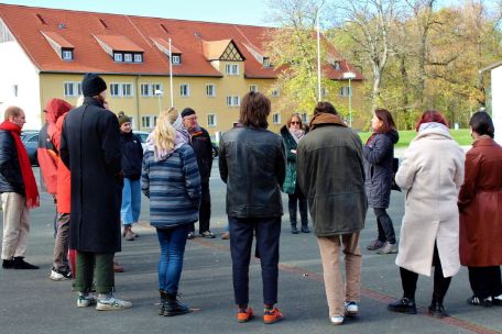 Eine Gruppe steht auf dem Parkplatz. Die Gruppe hört einer Referentin zu. Im Hintergrund ein Haus der internationalen Bildungssstätte.