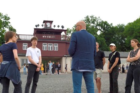 Ein Mitarbeiter der Gedenkstätte während eine Gruppe junger Menschen ihm zuhören. Im Hintergrund sind andere Gruppen. Noch weiter im Hintergrund sieht man das charakteristische Lagertor des ehemaligen Konzentrationslagers.