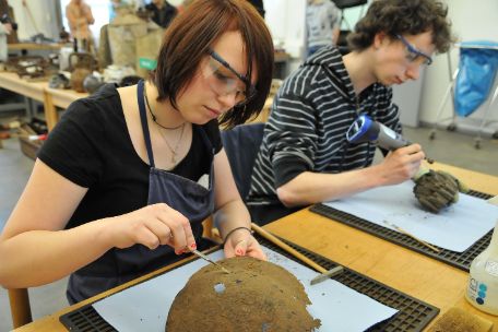 Zwei Teilnemer des Fundstückworkshops befreien zwei Fundstücke von Schmutz. Die junge Frau im Vordergrund bearbeitet augescheinlich eine Schüssel oder einen Helm, während der junge Mann hinter ihr ein Rohrteil bearbeitet.