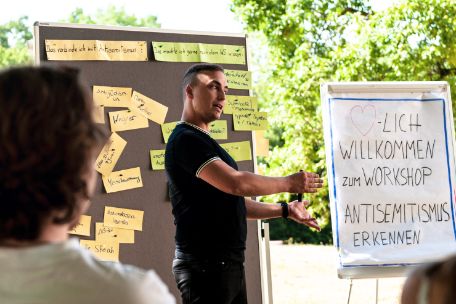 Ein Mitarbeiter der Bildungsabteilung steht im Freien neben einem Whiteboard mit der Aufschrift: "Willkommen zum Workshop: Antisemitismus Erkennen"